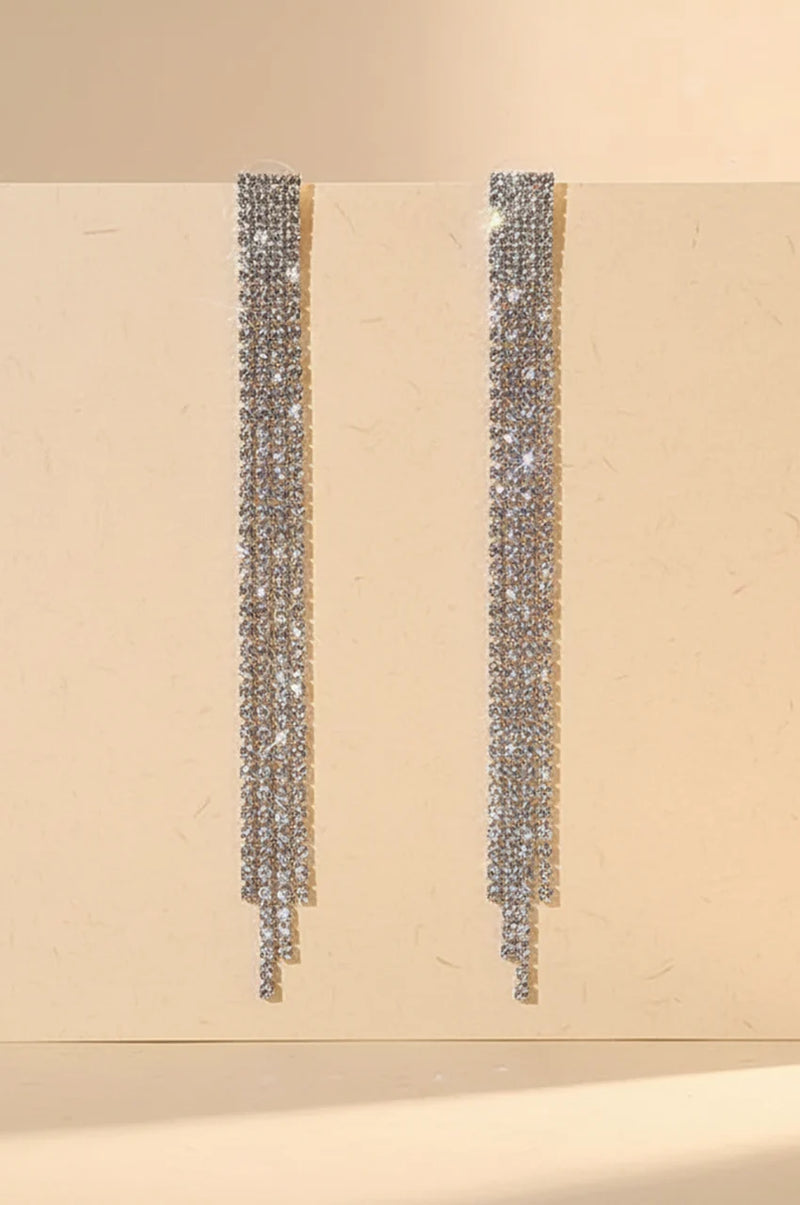 Front view of long silver rhinestone tassel earrings hanging against a beige background.