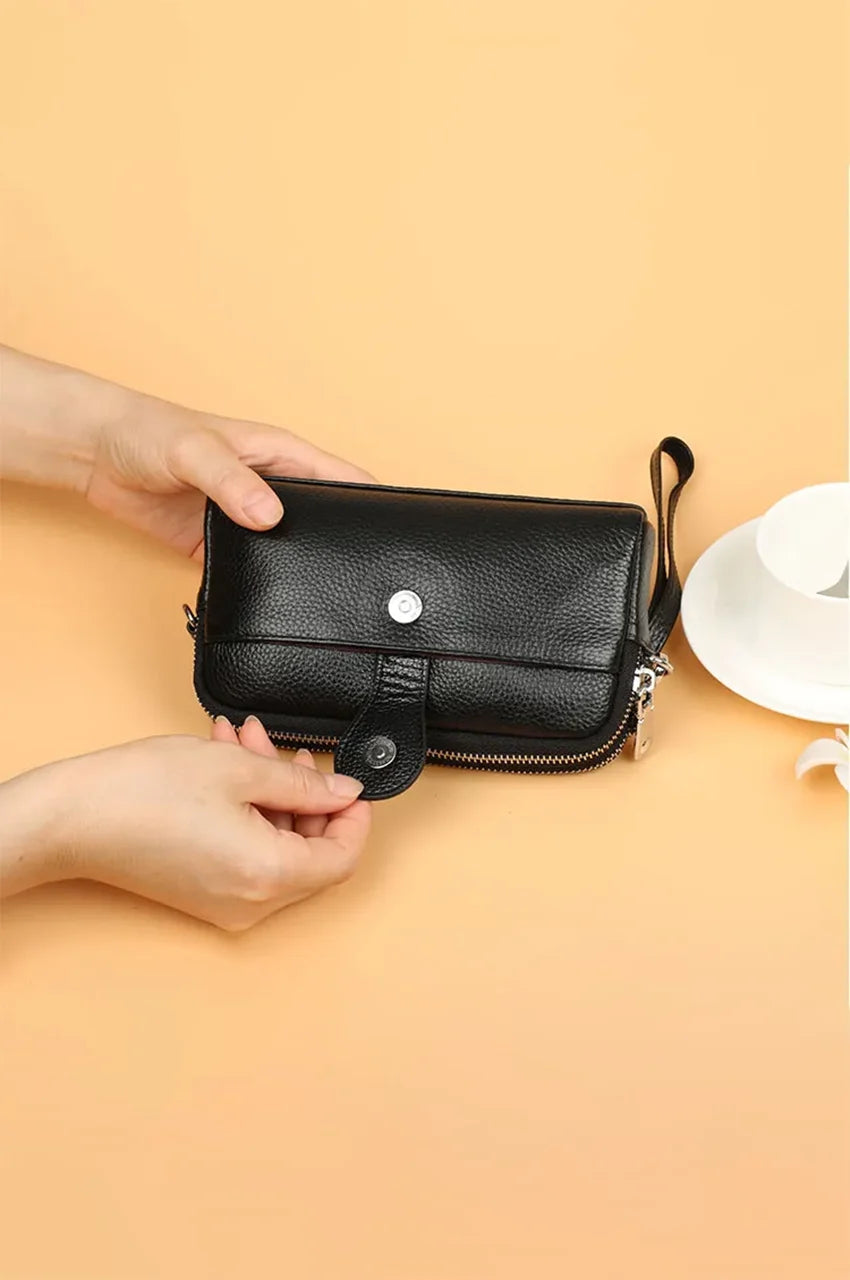 Hands opening the magnetic snap button tab on the front phone pocket of the black leather clutch.