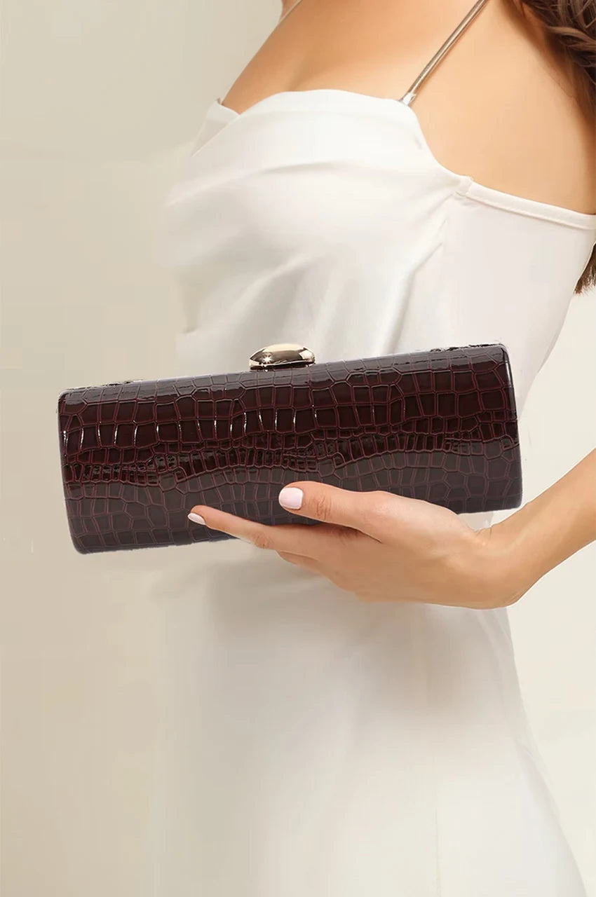 Woman holding a burgundy patent hard-shell clutch with gold details, styled for an evening wedding.