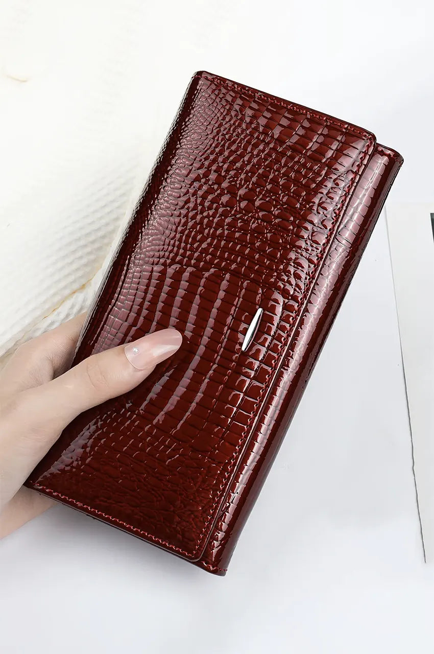 Hand holding the deep burgundy patent leather wallet clutch showing its glossy crocodile embossed finish.
