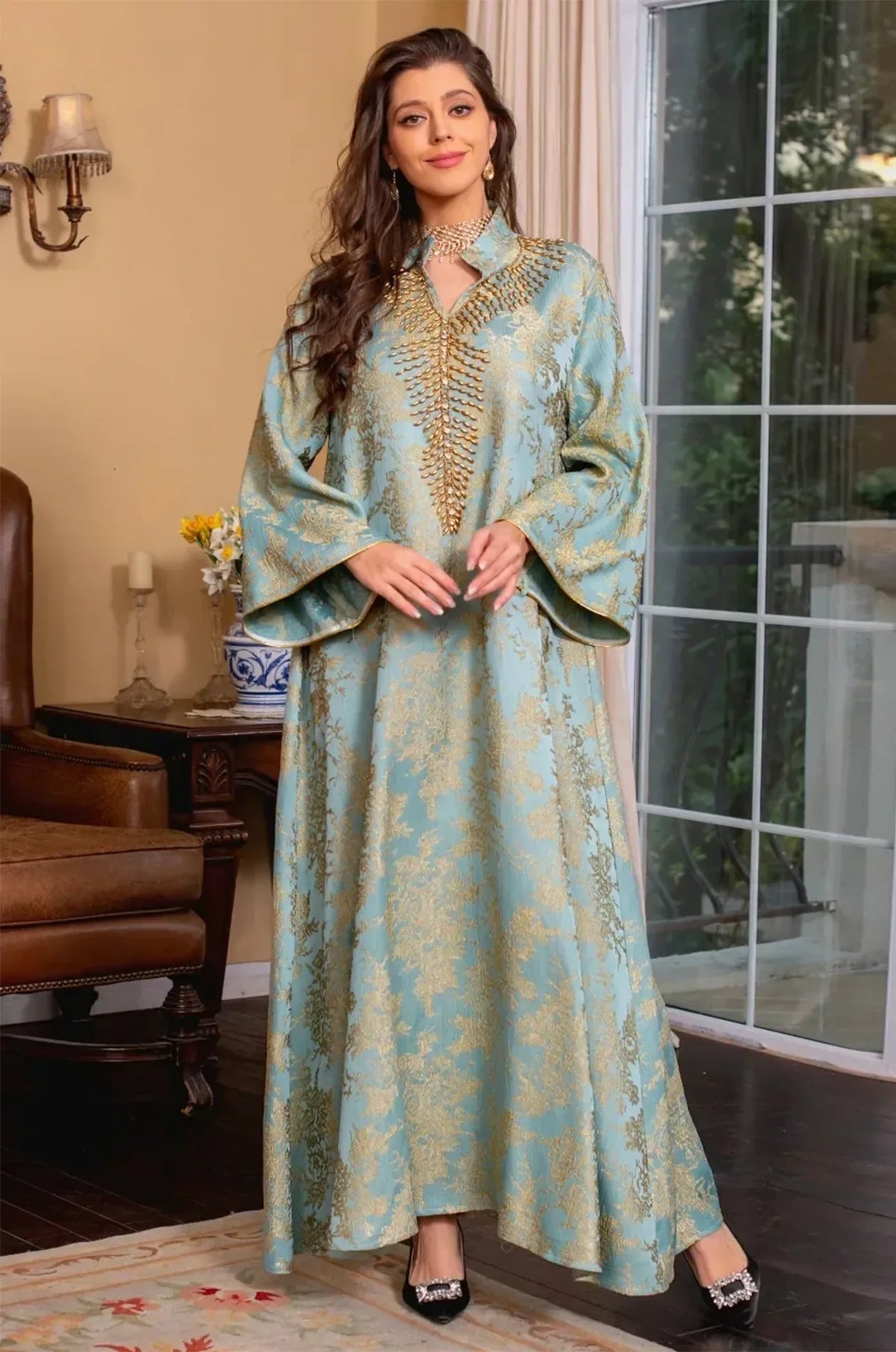 Full-body lifestyle shot of the Estel light blue kaftan standing in a luxurious room with a fireplace and vintage decor.