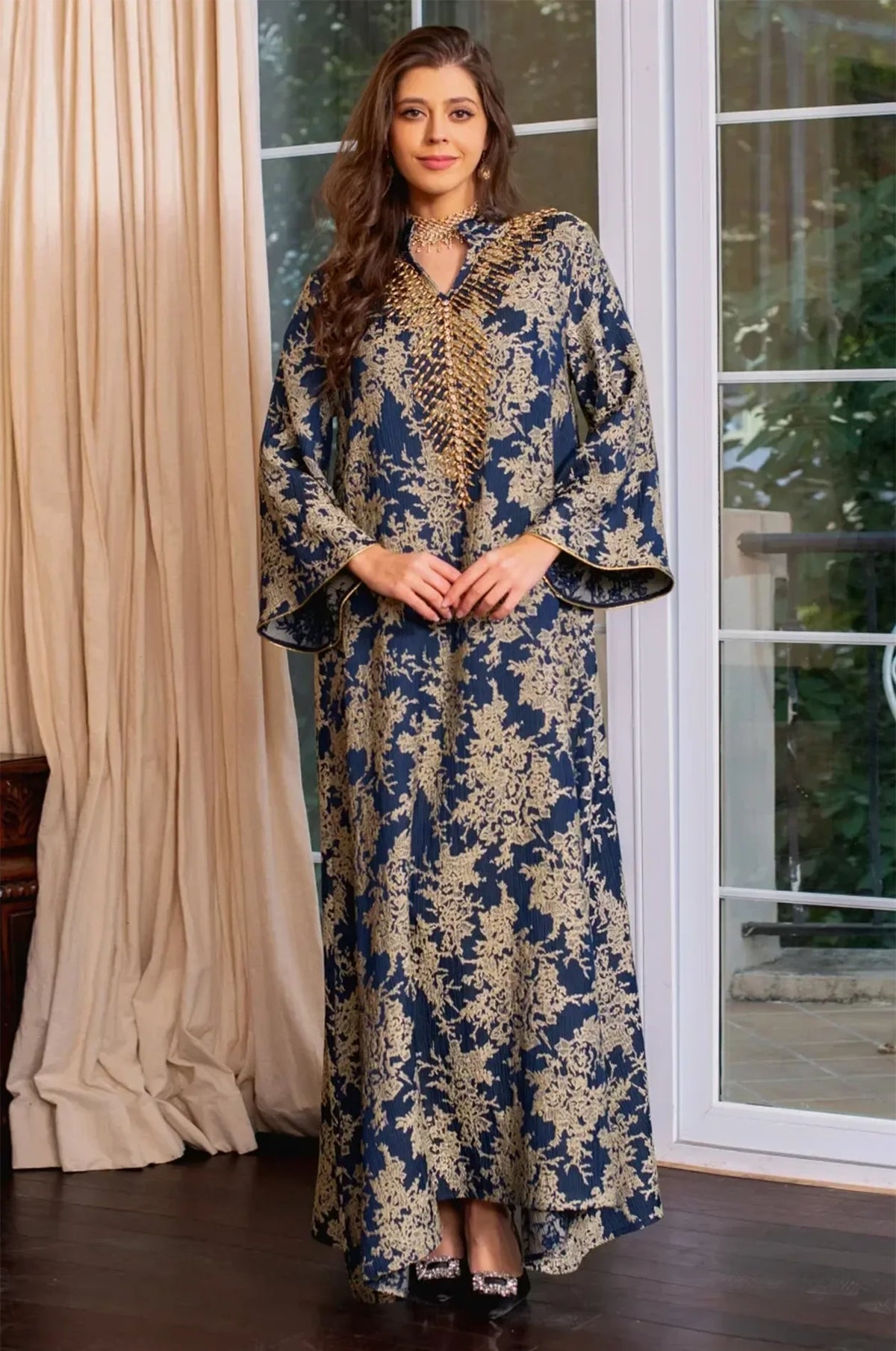 Full-length view of the Estel navy blue kaftan with hands clasped, posed in front of white French doors and curtains.