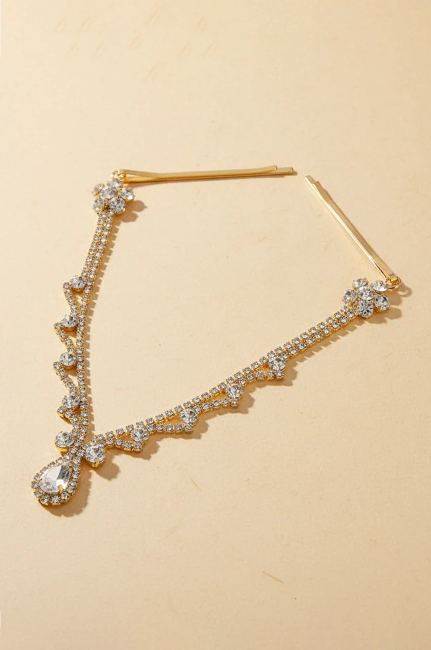 Full view of gold rhinestone forehead headpiece with teardrop crystal and hair pins on a beige background.
