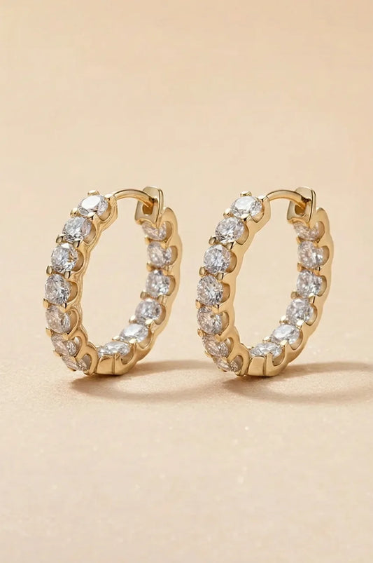 Pair of yellow gold huggie earrings with round cut moissanite stones on a beige background.