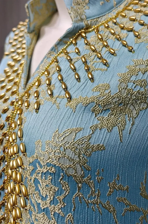 Macro close-up shot of the light blue textured jacquard fabric and hand-applied gold teardrop beads on the Estel kaftan.