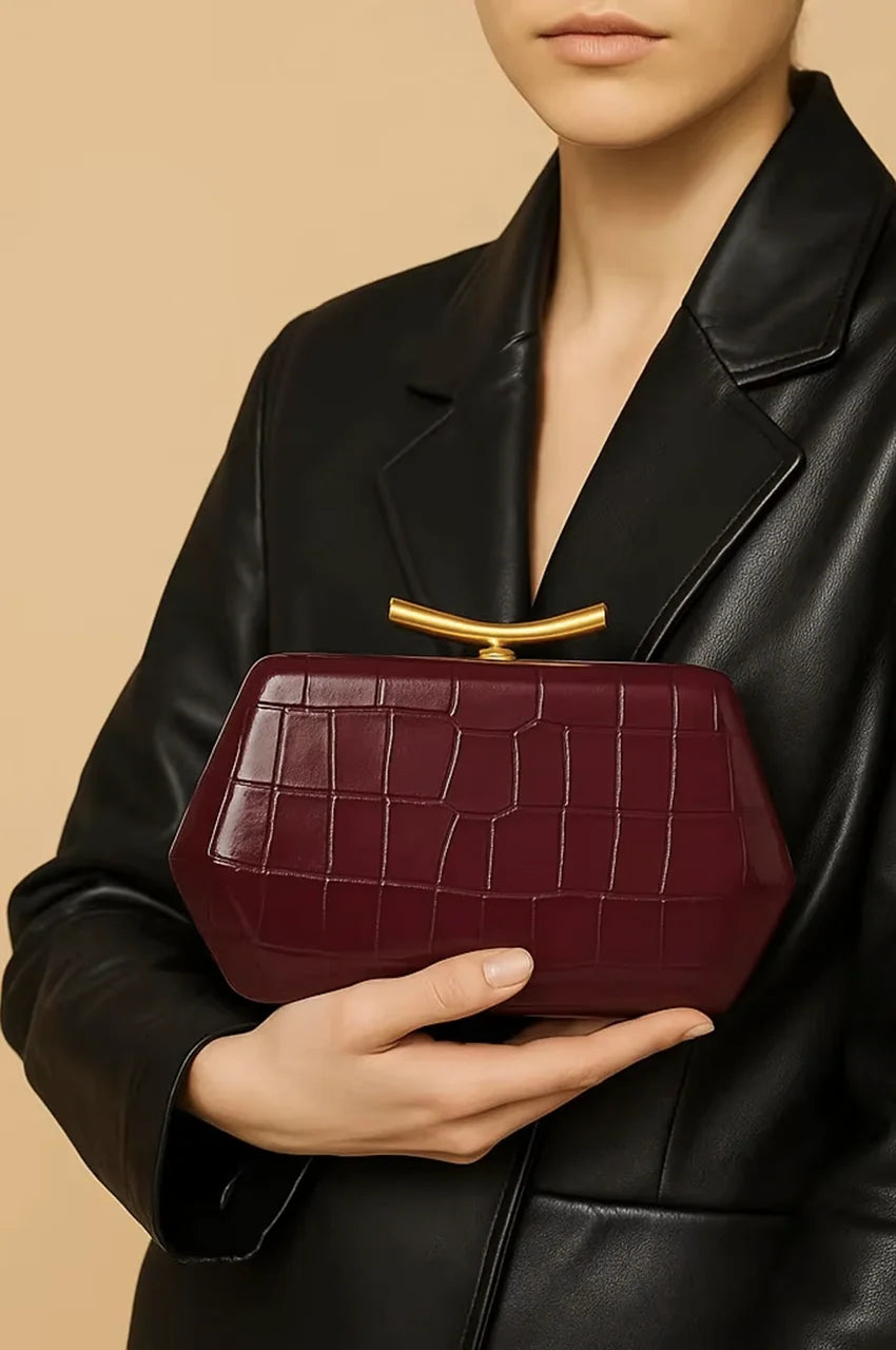 Woman holding a burgundy patent crocodile-embossed clutch bag against a black leather blazer.