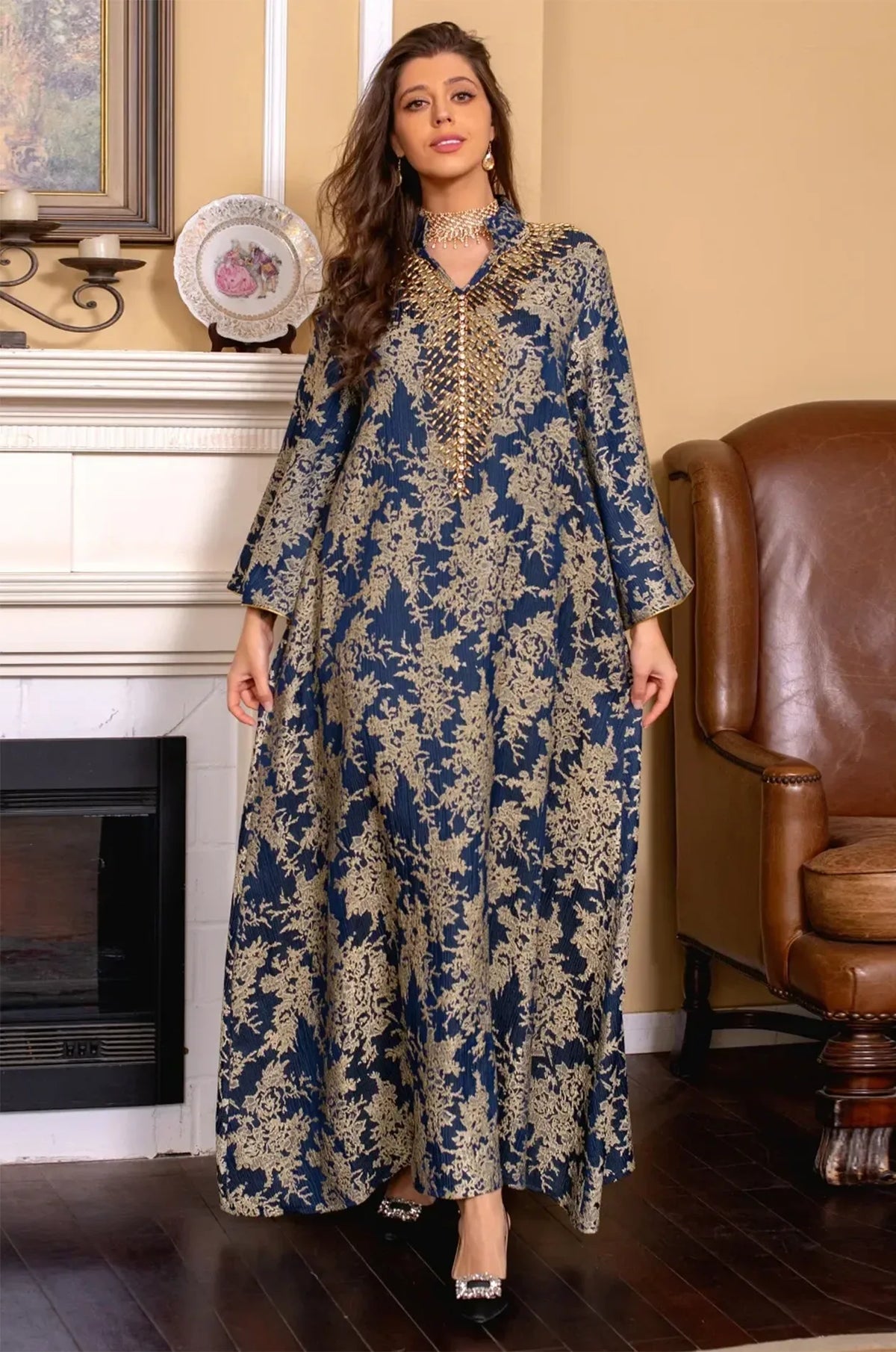 Lifestyle shot of the Estel navy blue kaftan with the model standing in a luxury interior near a classic white fireplace.