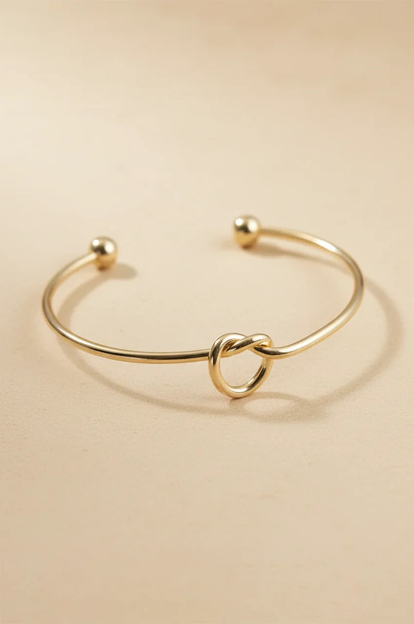 Minimalist gold open cuff bracelet with love knot detail on beige background.
