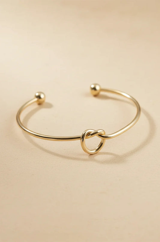 Minimalist gold open cuff bracelet with love knot detail on beige background.