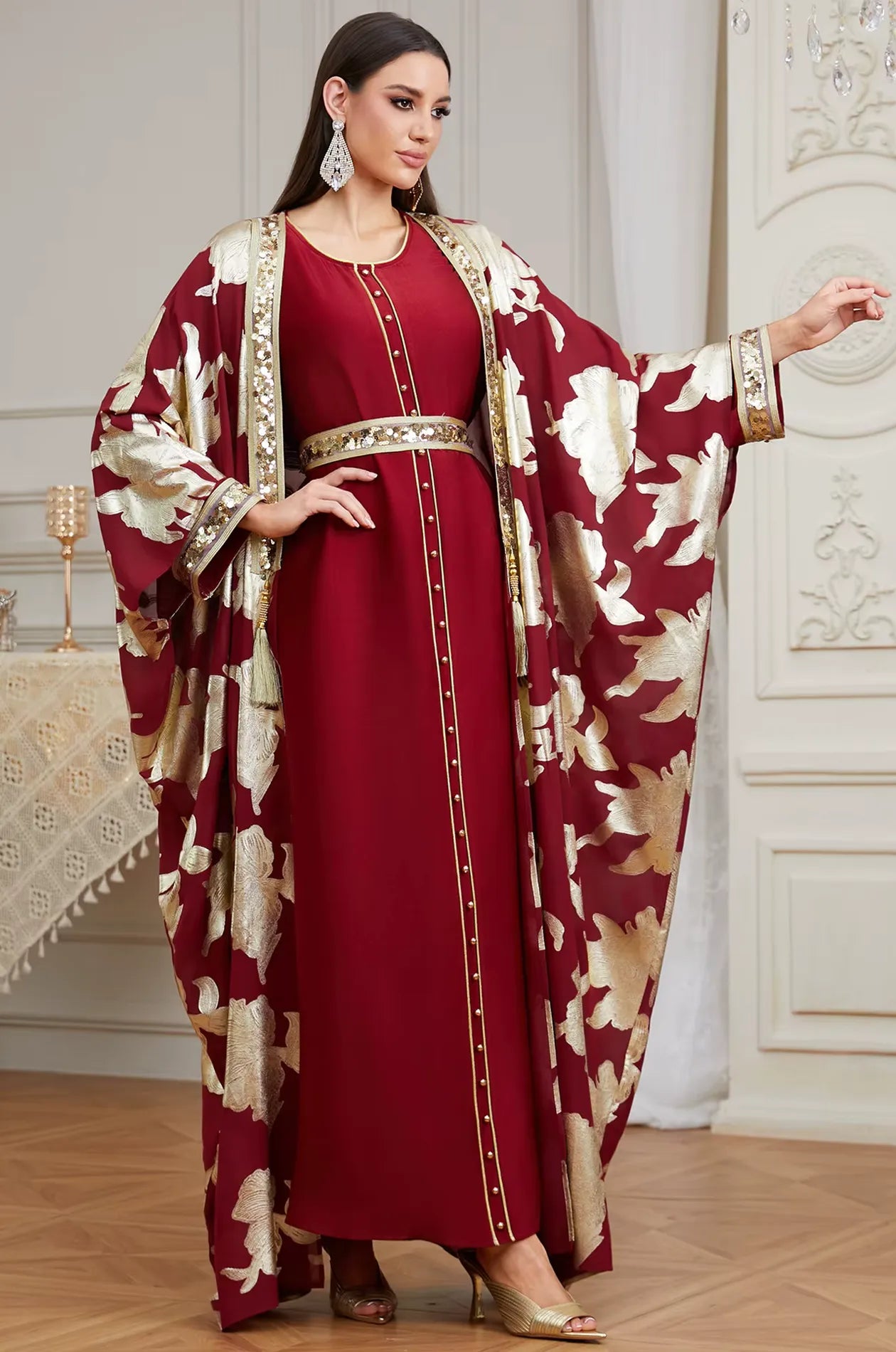 Full-length shot of the red Puppis kaftan luxe with arms extended to display the majestic cape-style design and gold floral accents.
