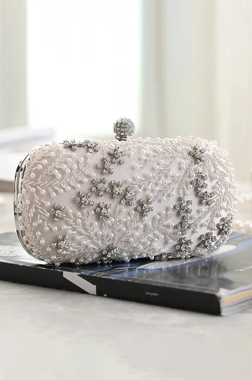 Side profile of white luxury evening bag resting on a surface, highlighting the 3D texture of the floral beading and structured oval shape.