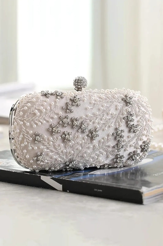 Side profile of white luxury evening bag resting on a surface, highlighting the 3D texture of the floral beading and structured oval shape.