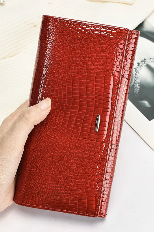 Hand holding the vibrant red patent leather wallet clutch showing its glossy crocodile texture.